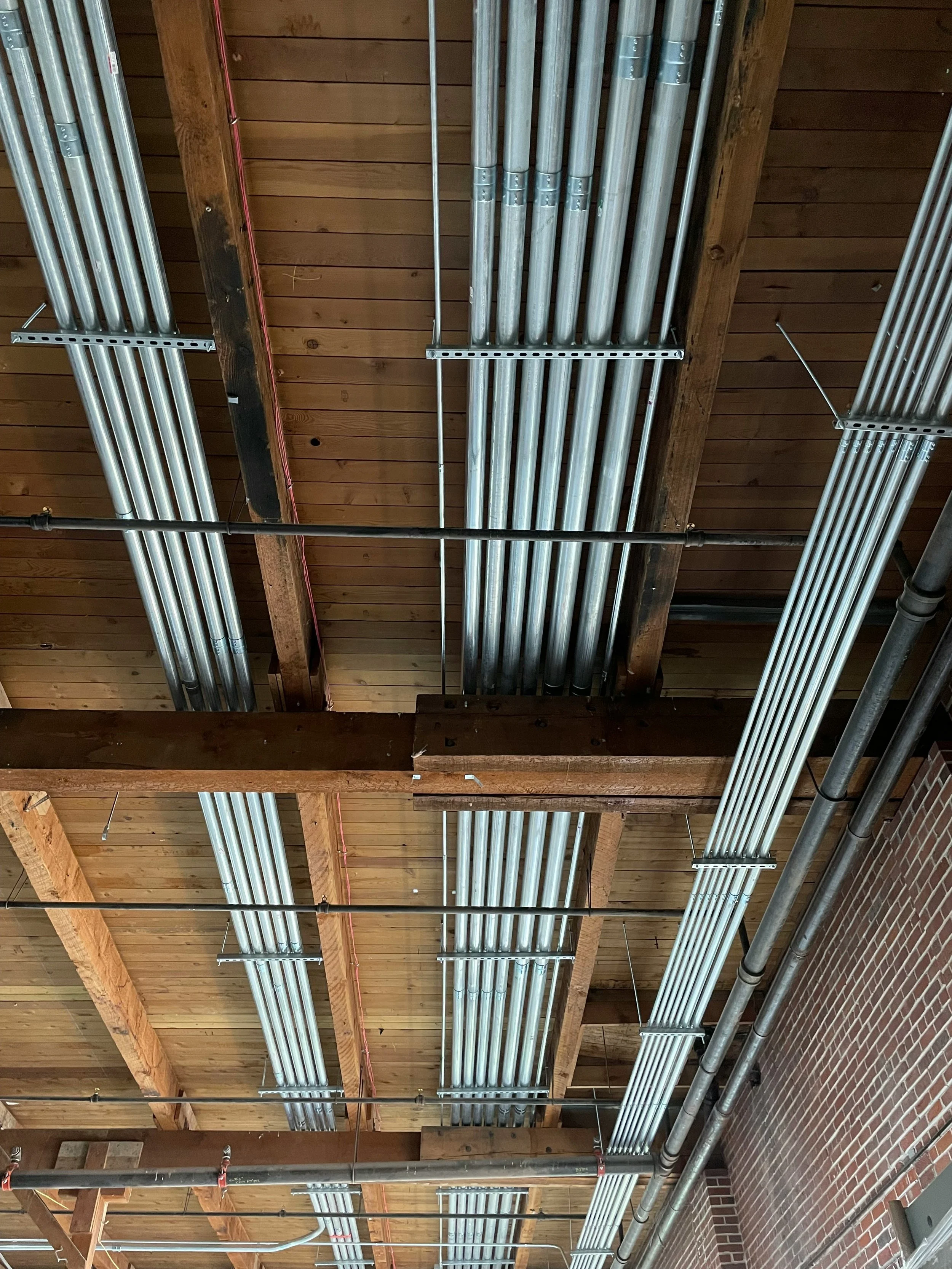 Ceiling with exposed wooden beams, metal conduit pipes, and hanging metal cable trays in a building under construction.