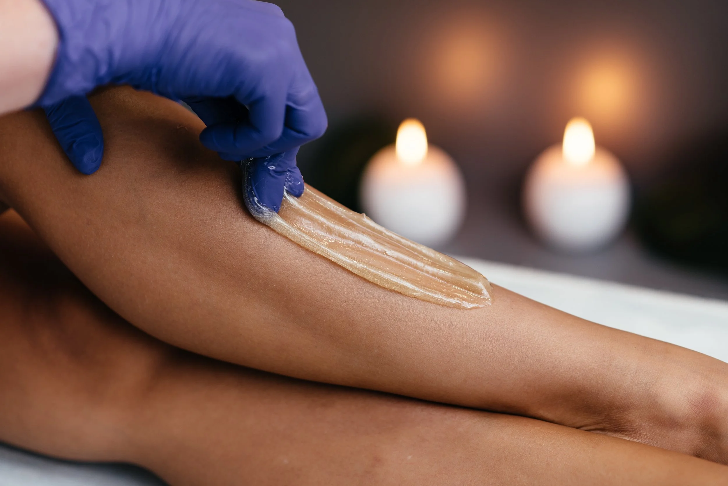 A person with a gloved hand applies sugar paste to the leg of a client for hair removal.