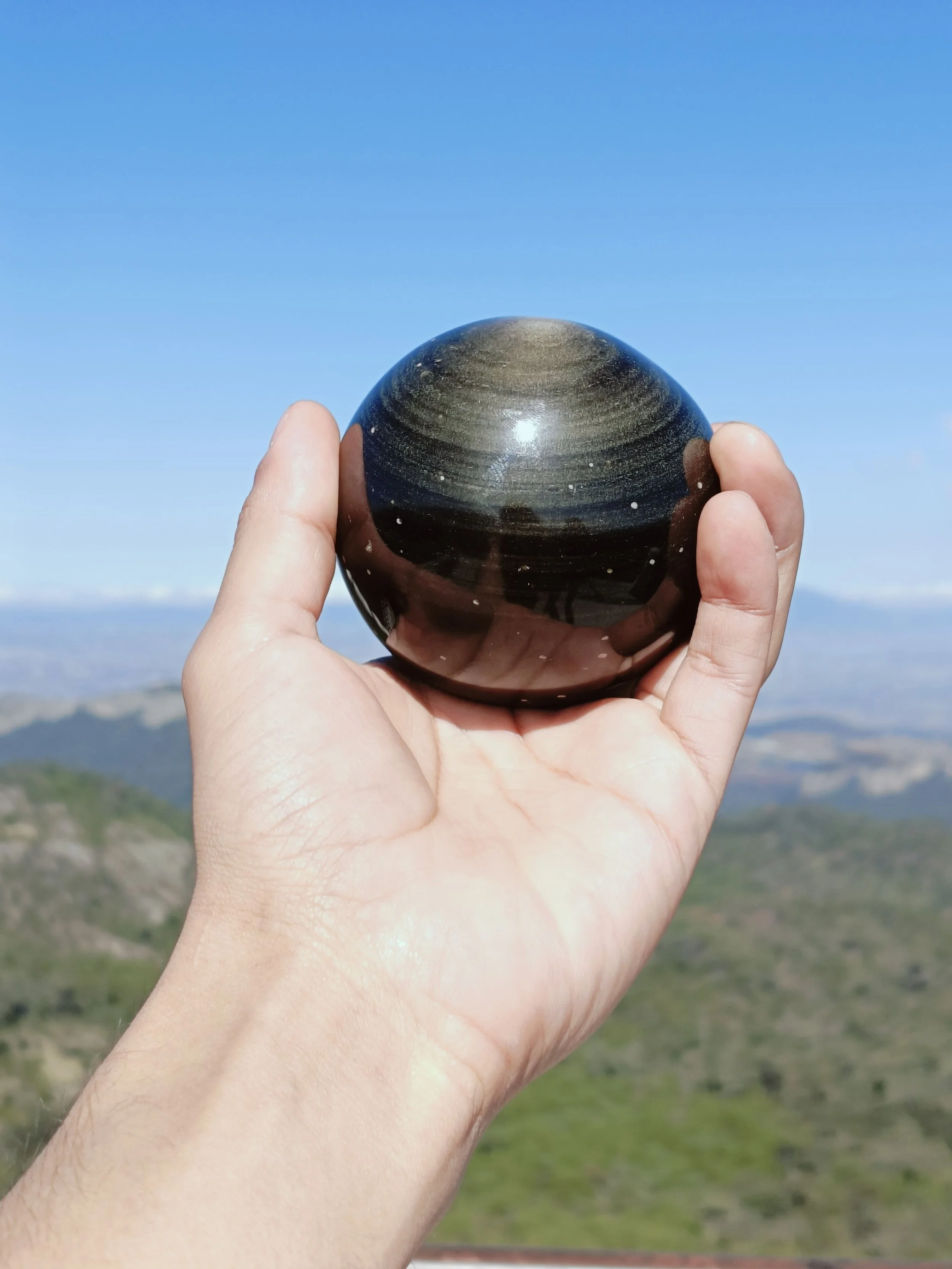 Mano sosteniendo una piedra negra y redonda con un paisaje de montañas y cielo azul al fondo.
