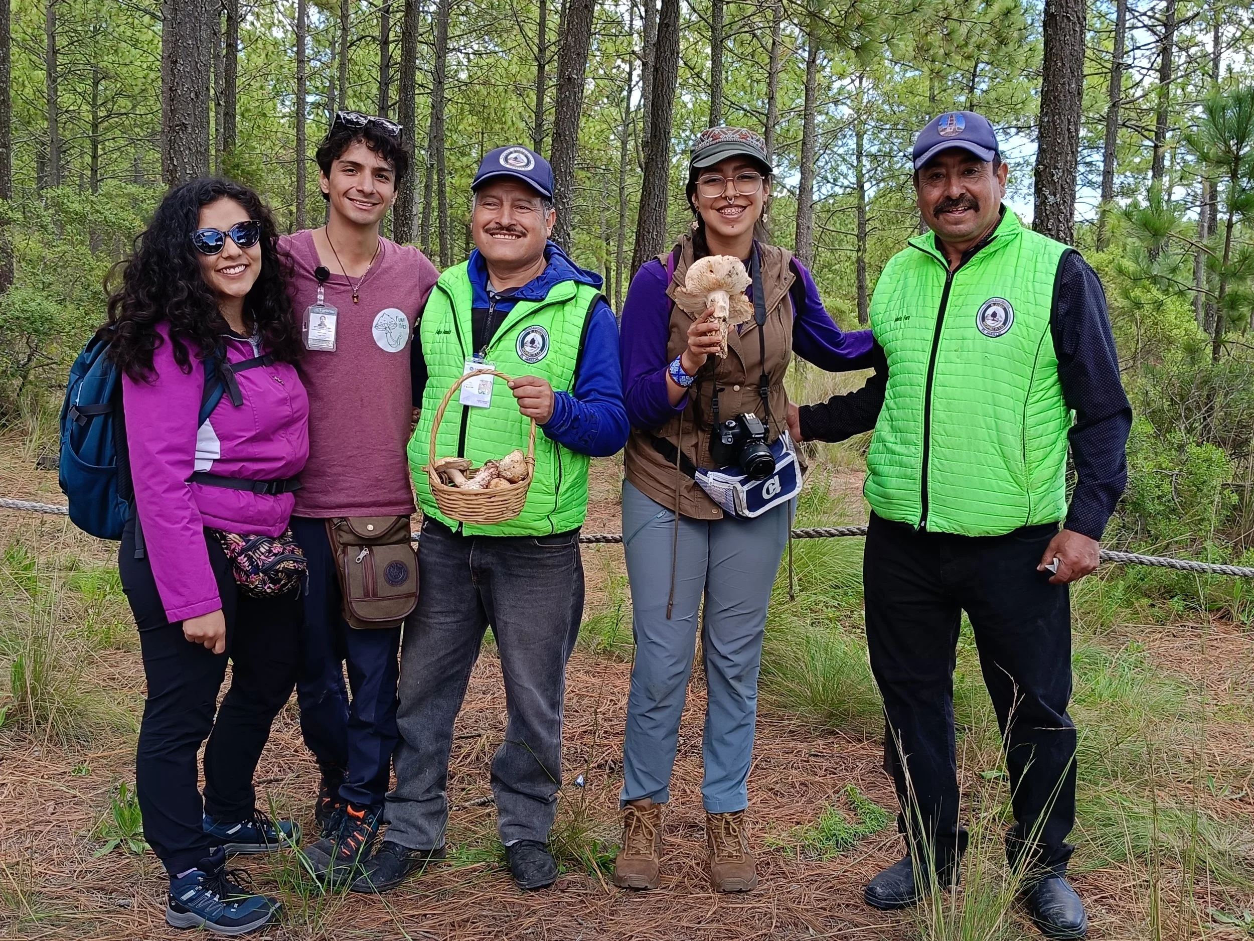 Grupo de seis personas en un bosque, sonriendo y posando para la foto, algunos con equipamiento de senderismo y dos sosteniendo hongos
