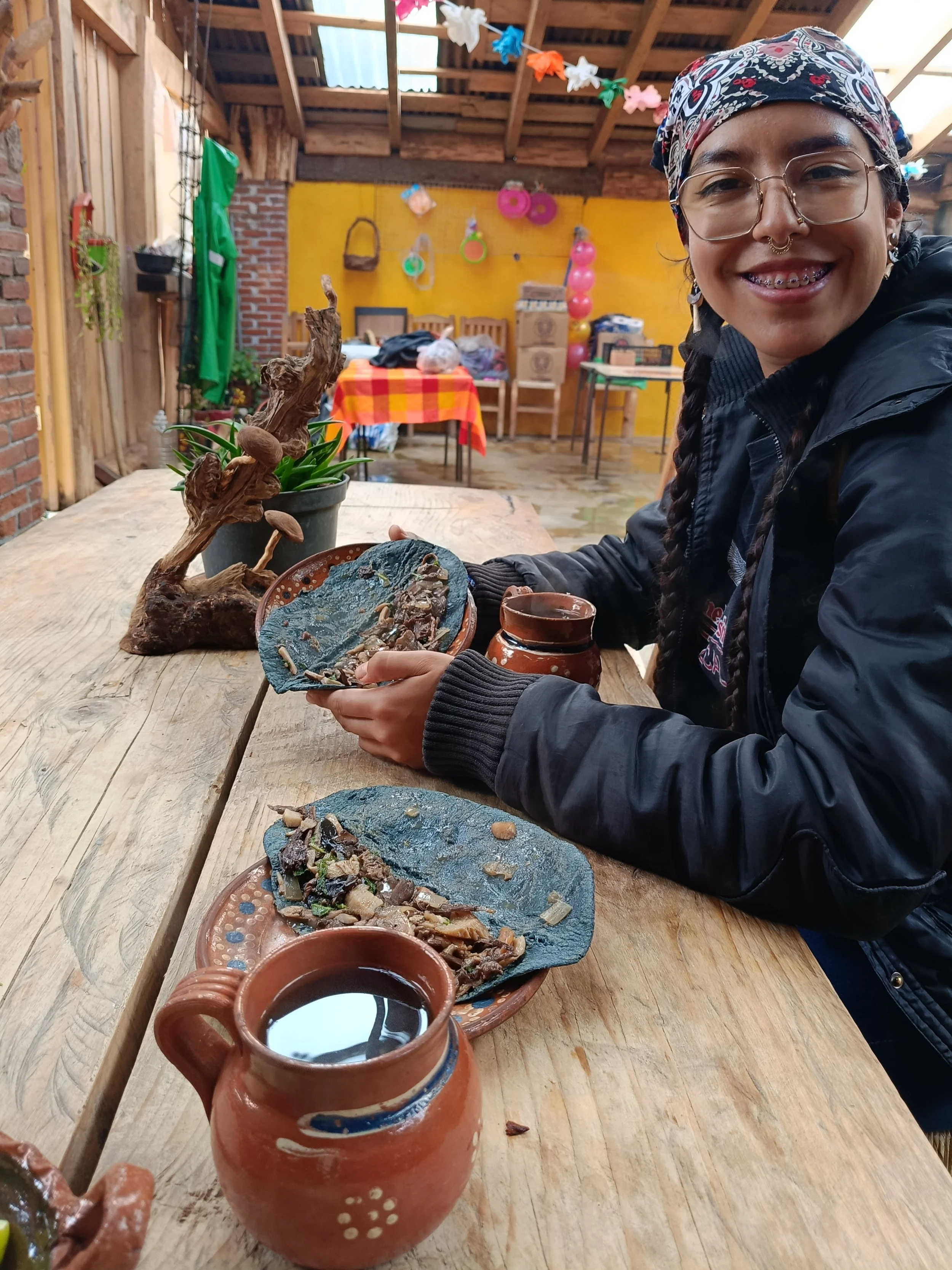 Una mujer sonriente, con gafas y un pañuelo en la cabeza, sentada en una mesa de madera en un espacio decorado con globos y adornos coloridos, disfrutando de comida tradicional mexicana, con platos de piedra y una jarra de barro con bebida.