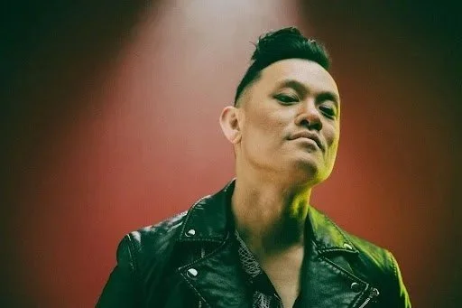 Portrait of a person with a confident expression, wearing a black leather jacket, with a short mohawk hairstyle, and slightly raised chin, against a dark background with red and green lighting. Photo Credit by Tadin Brego of Blacklodge Portland
