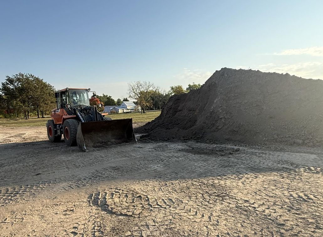 Construction site with a front loader next to a large pile of dark dirt or soil, on a partly cloudy day with trees and buildings in the background.