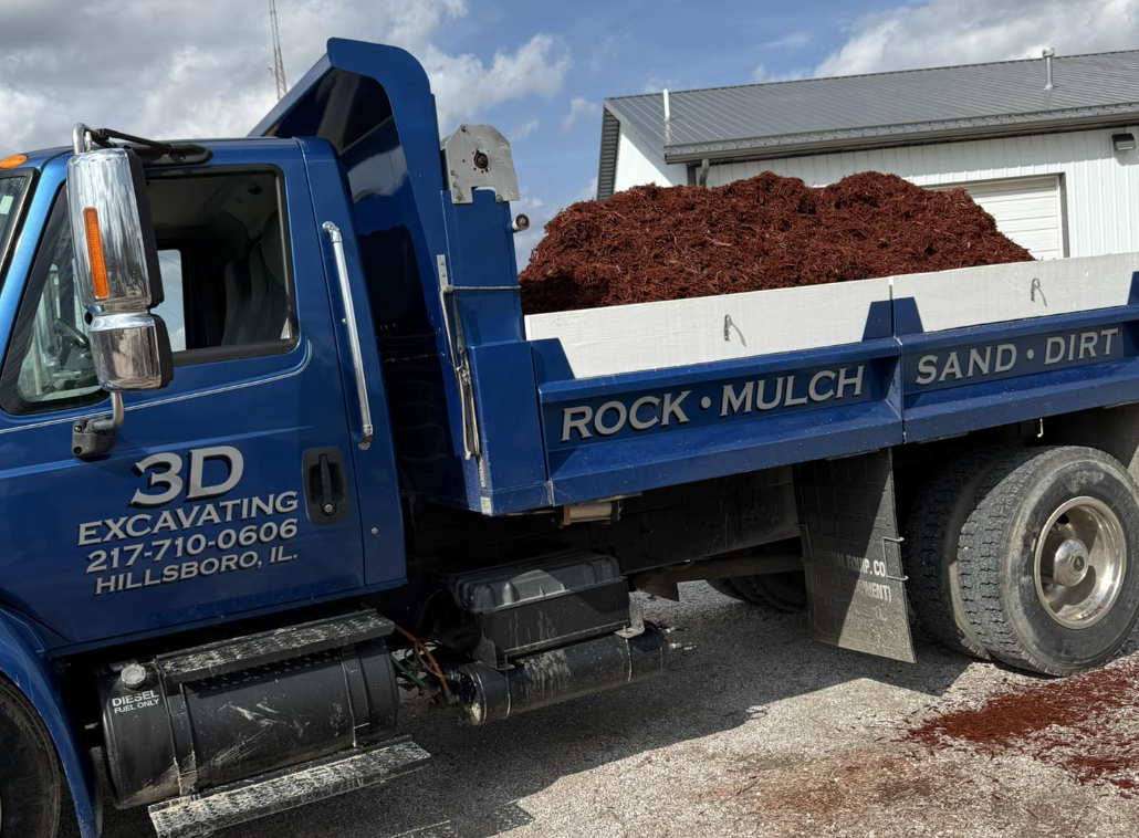 Blue dump truck filled with red mulch parked on a gravel surface near a white industrial building with a gray metal roof.