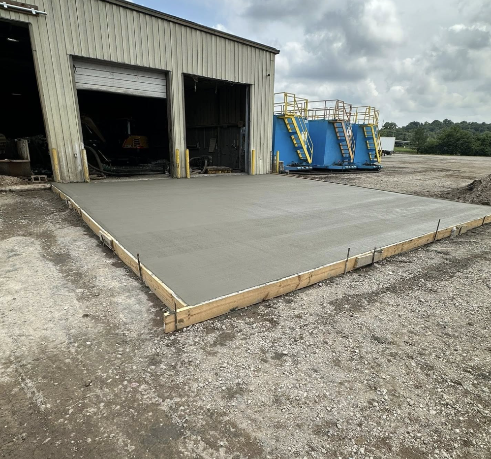 Freshly poured concrete slab outside a metal building with open garage doors, with blue tanks and construction equipment in the background.