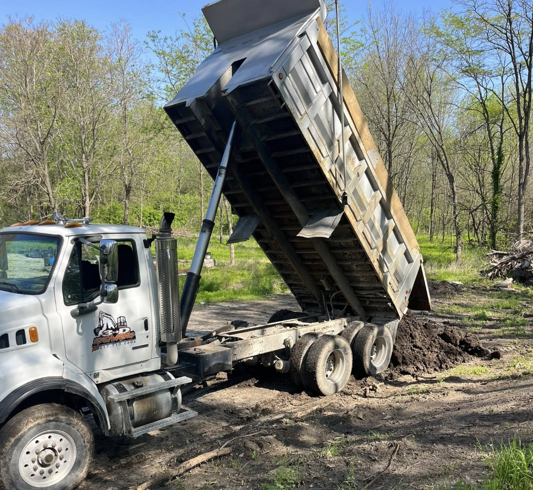 A dump truck is dumping dirt or soil onto the ground in a wooded outdoor area with trees and blue sky in the background.