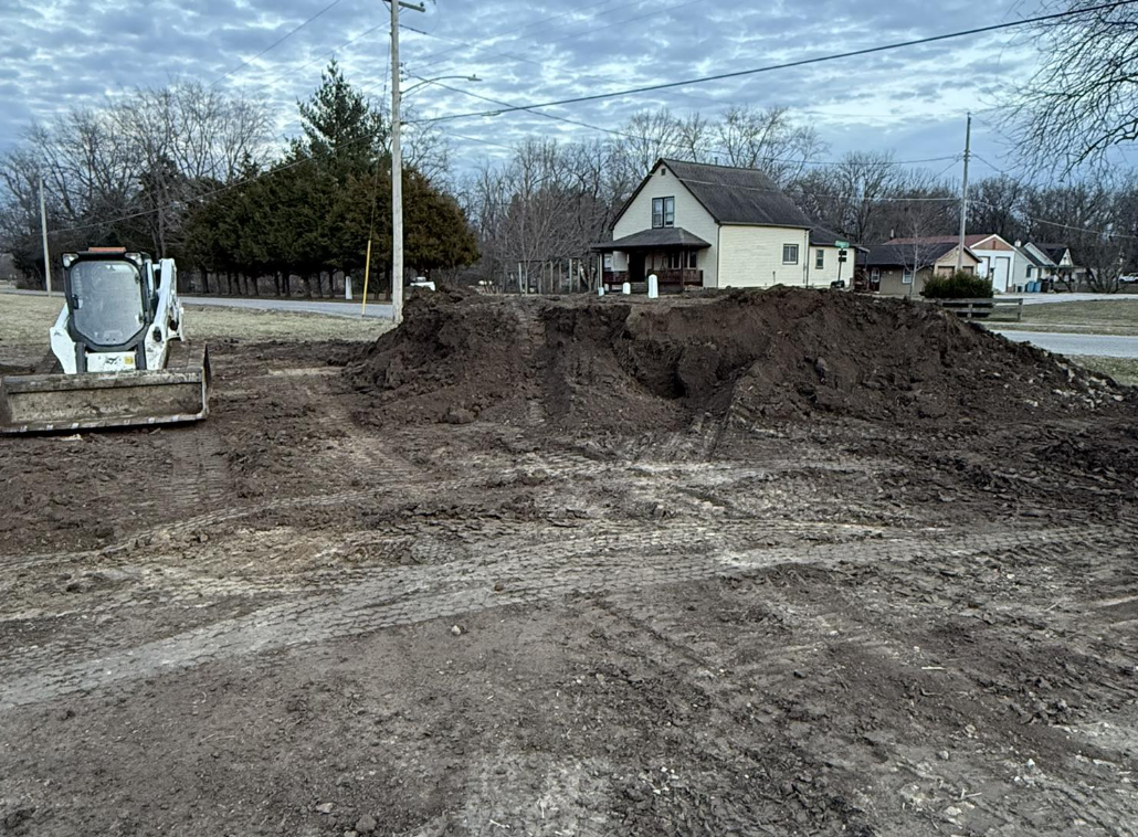 Construction site with dirt piles and a small skid steer loader, residential houses in the background under a cloudy sky.
