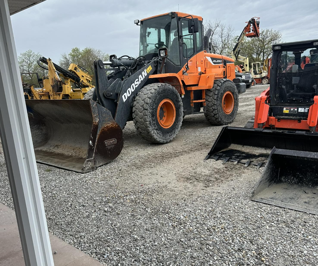 Orange Doosan wheel loader parked on gravel, with a black bucket attachment, surrounded by other construction equipment under a cloudy sky.