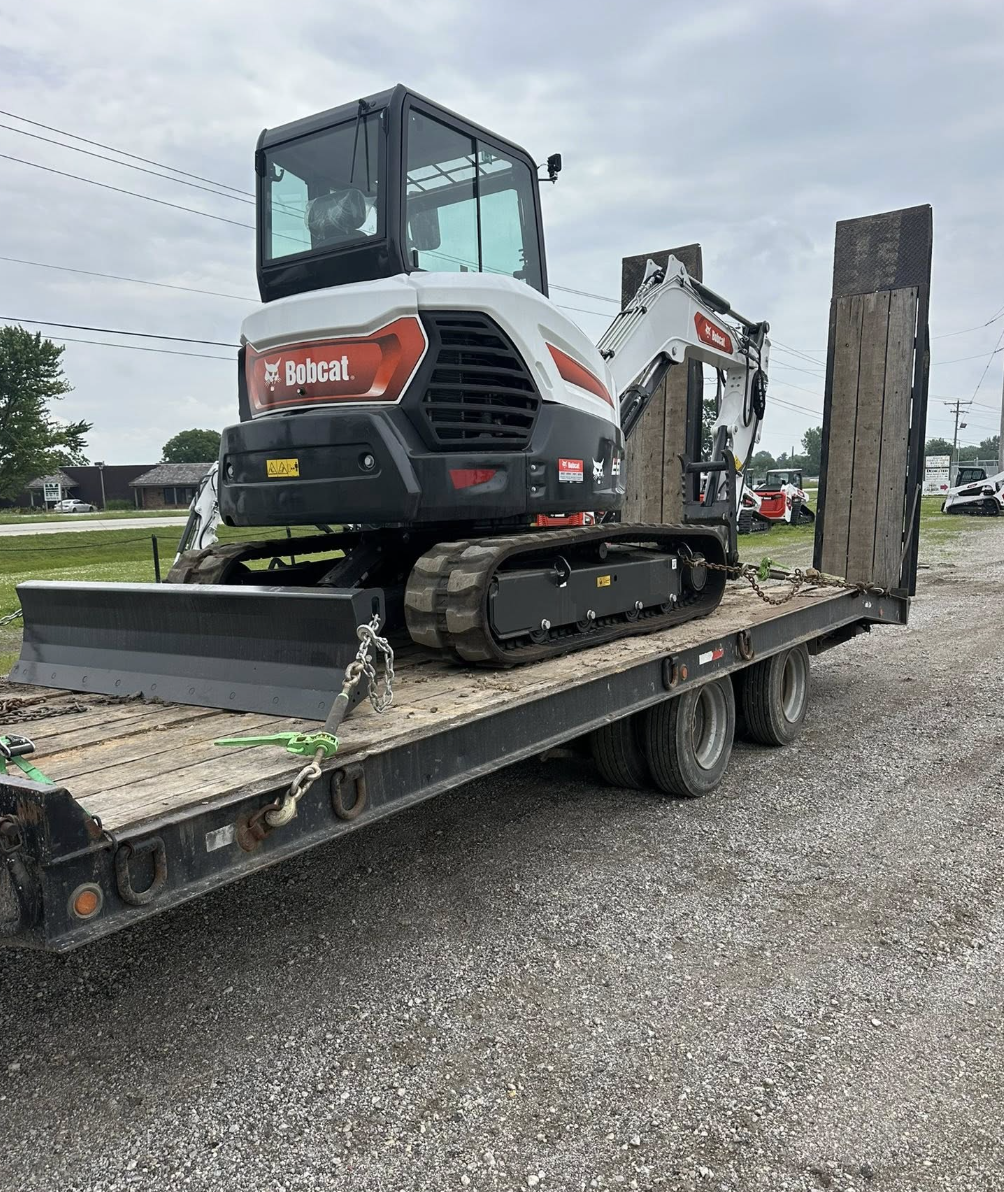 A Bobcat mini excavator loaded onto a flatbed trailer, secured with chains, against an outdoor background with trees, power lines, and parked vehicles.
