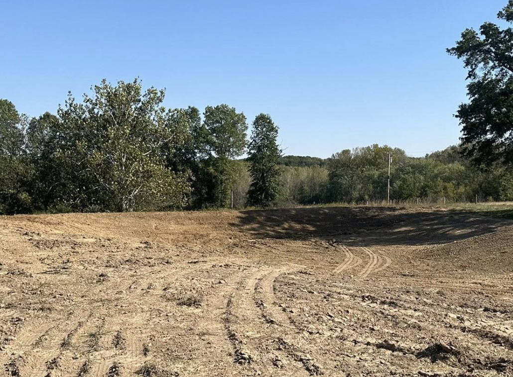 A dirt clearing with tire tracks, surrounded by trees and a clear blue sky.