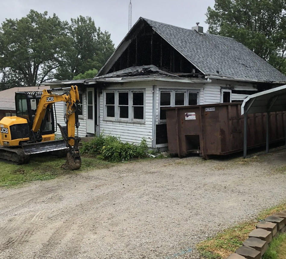 A damaged house with a partially collapsed roof, boarded-up windows, a small yellow JCB excavator parked outside, and a large rusty dumpster nearby.