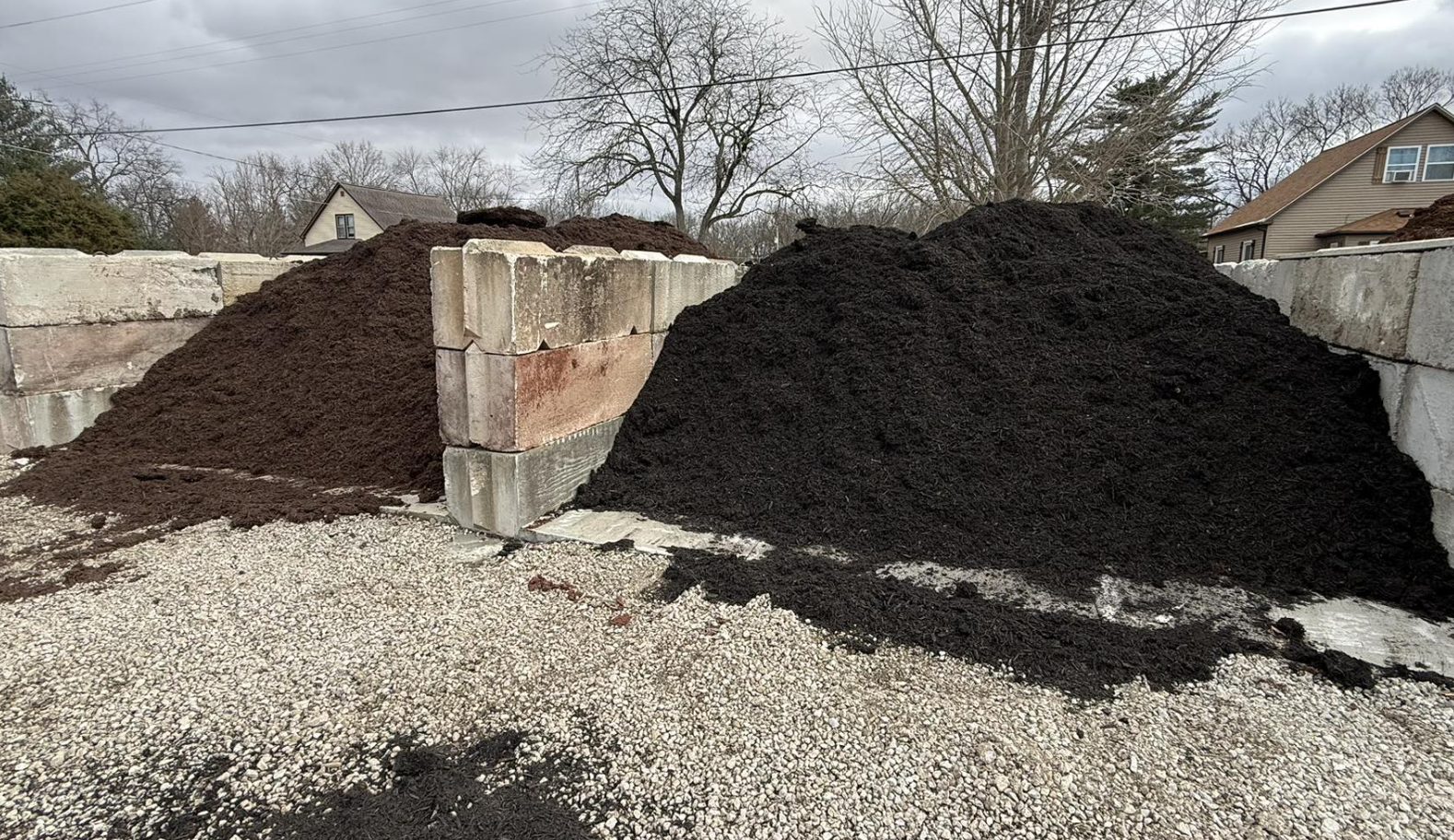 Two piles of dark soil or mulch on a construction site, with a brick and concrete wall in the background and cloudy sky overhead.