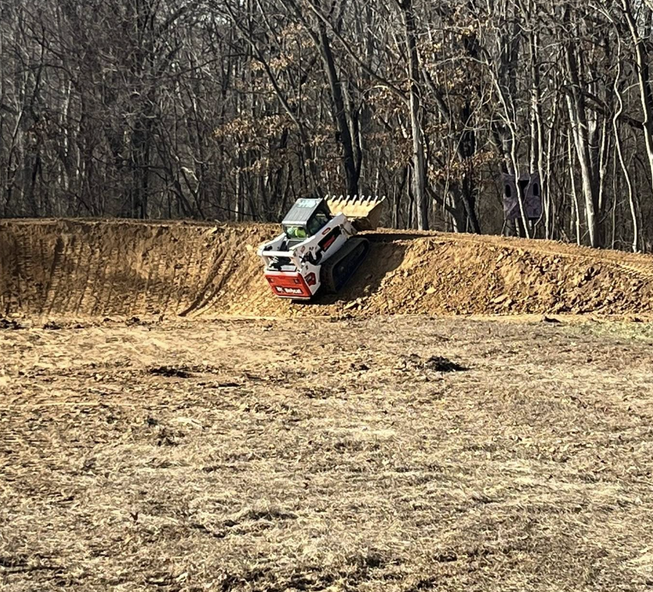 A small remote-controlled excavator on a dirt mound in a wooded area, with barren trees and a purple object in the background.