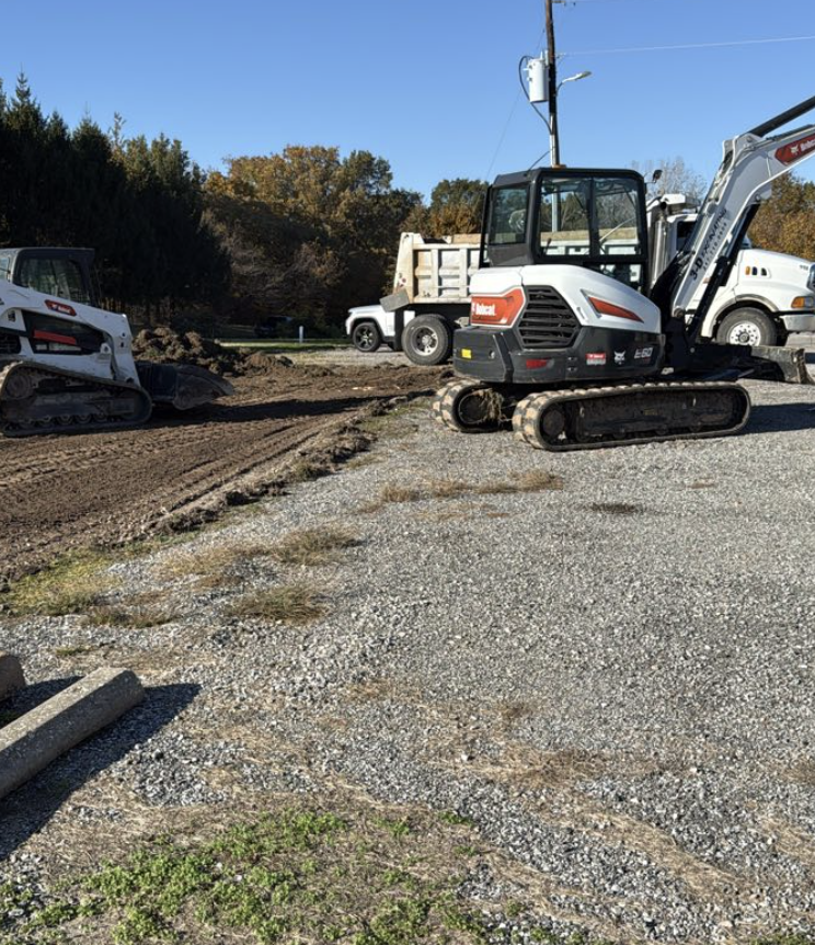 Construction site with small excavator and compact track loader on gravel and dirt area, trees with fall foliage in background, and a utility pole with power lines.