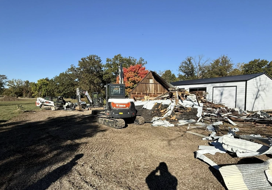Debris from a destroyed building, excavation equipment on a dirt lot, clear blue sky, and some trees in the background.