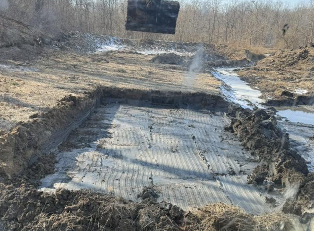A construction site with a large excavated area and a reflectively lined base, surrounded by dirt and mud, with some water and a wooded area in the background.