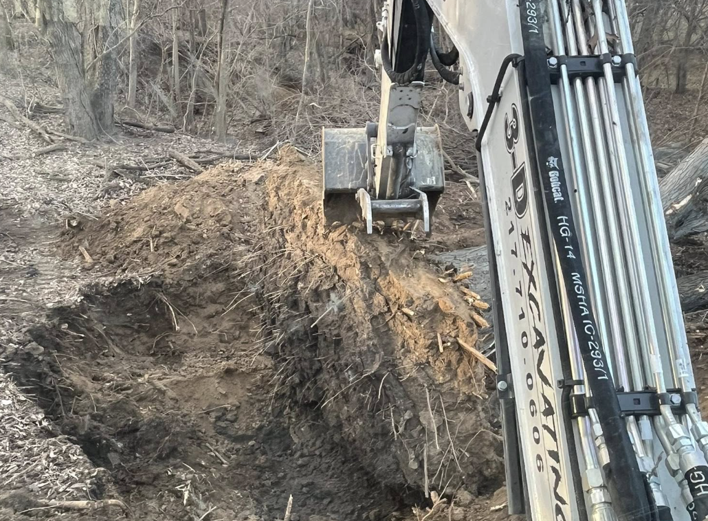 A hydraulic excavator digging into the ground in a wooded area.