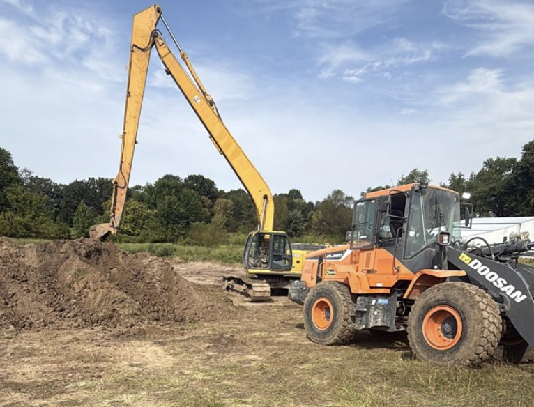 Construction site with an orange wheel loader and a yellow excavator digging earth, with trees and a blue sky in the background.