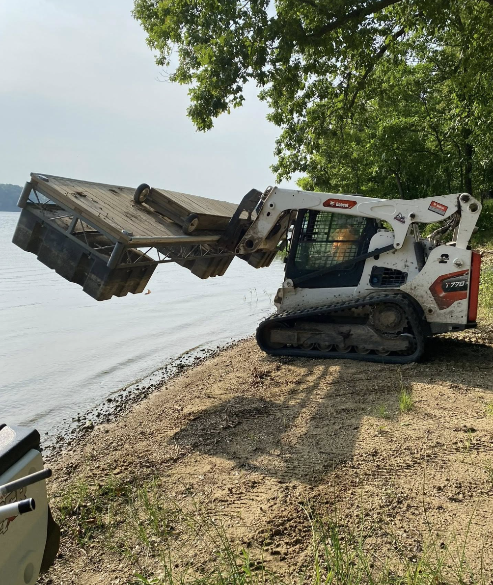 A small Bobcat T770 compact track loader with a grapple attachment lifting a wooden dock off the shoreline near kayaks and green trees.