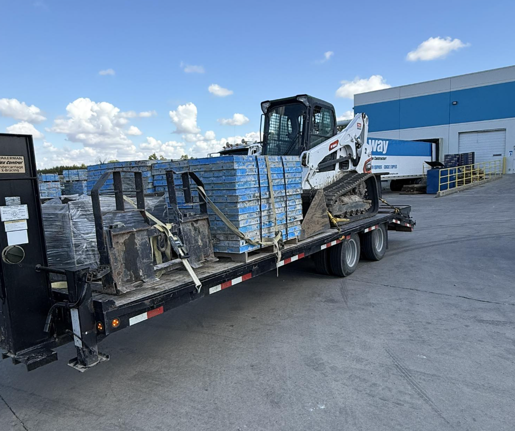 Flatbed truck transporting construction equipment, including a small crane and blue pallets, outside a warehouse under a partly cloudy sky.