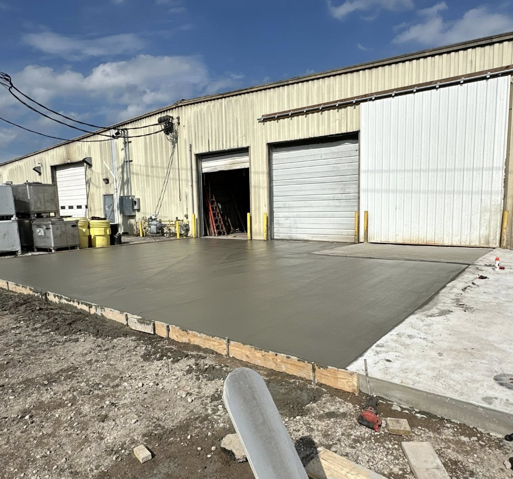 Concrete being poured and leveled in front of a warehouse with three garage doors, some construction equipment, and a clear sky.