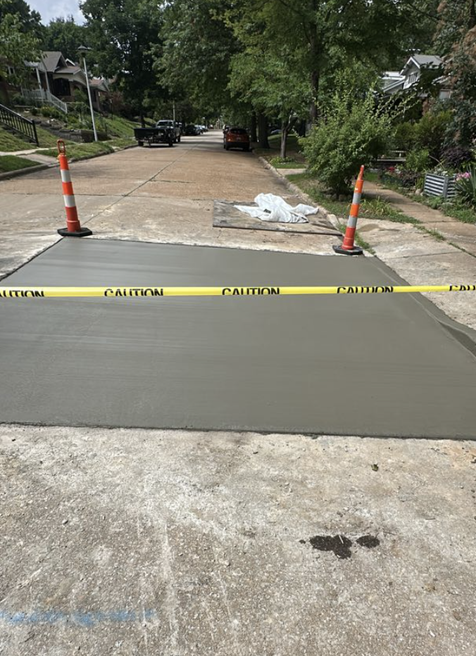 Fresh asphalt being laid on a residential street, sectioned off with caution tape and orange traffic cones, with weathered pavement and green trees lining the street.