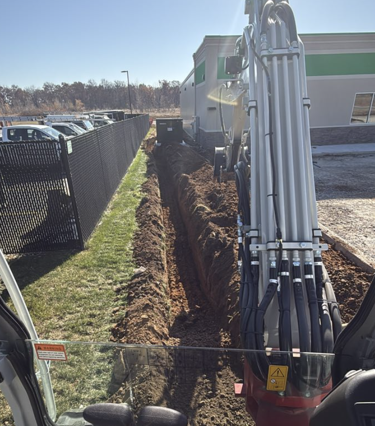 Construction equipment digging a trench parallel to a black metal fence near parked cars.