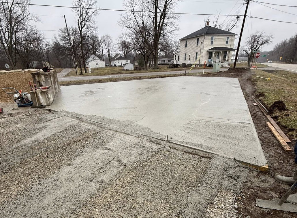 Freshly poured concrete slab on a construction site, with construction tools and materials around, and houses in the background.