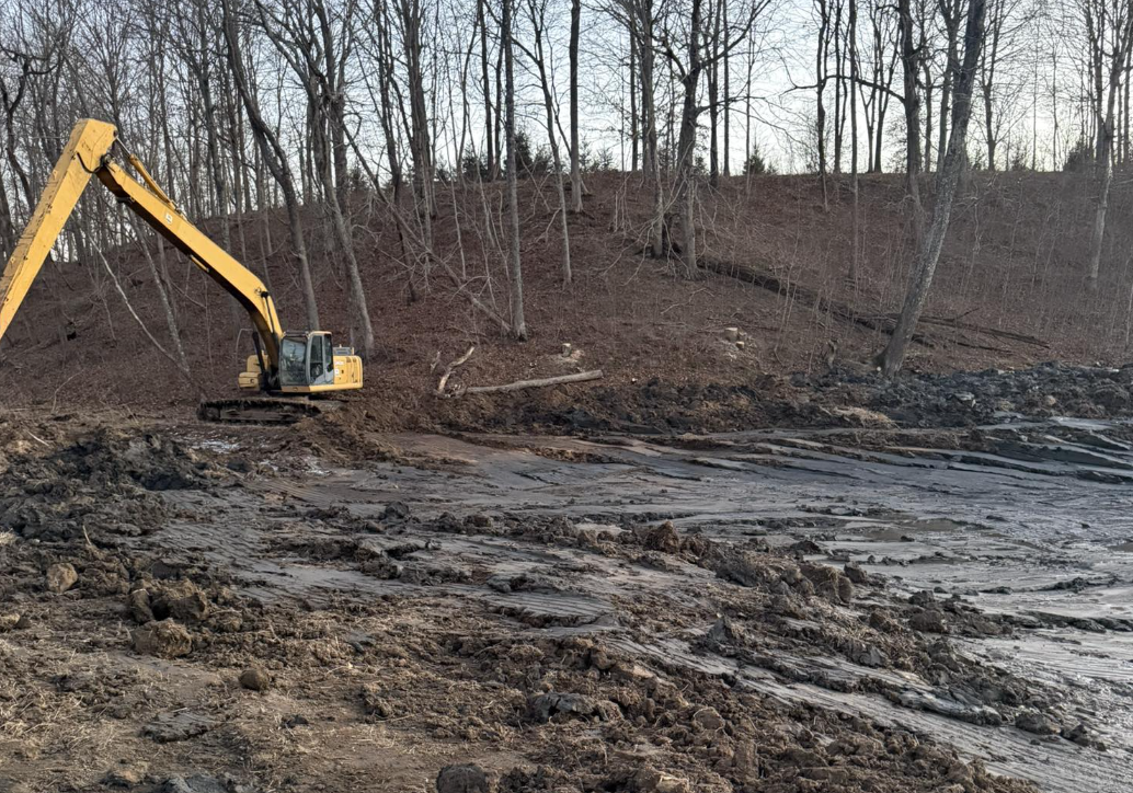 An excavator working on a muddy slope in a wooded area, clearing or excavating soil and debris.