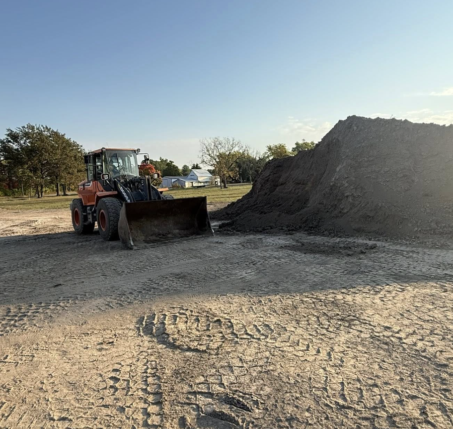 Construction site with a backhoe loader and a large pile of dirt on a dirt surface with tire tracks, trees, and buildings in the background under a blue sky.
