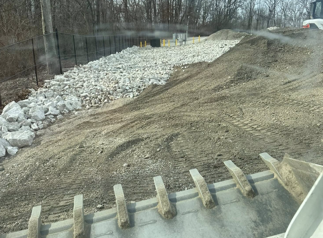 Construction site with gravel, dirt, and rocks, fenced off with a black mesh fence, and a white vehicle parked at the edge of the area.