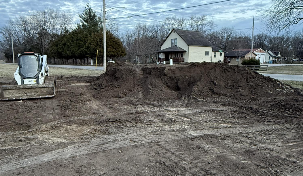 A construction site with a small skid steer loader on the left and a large pile of dirt in the center, with a residential neighborhood and trees in the background.