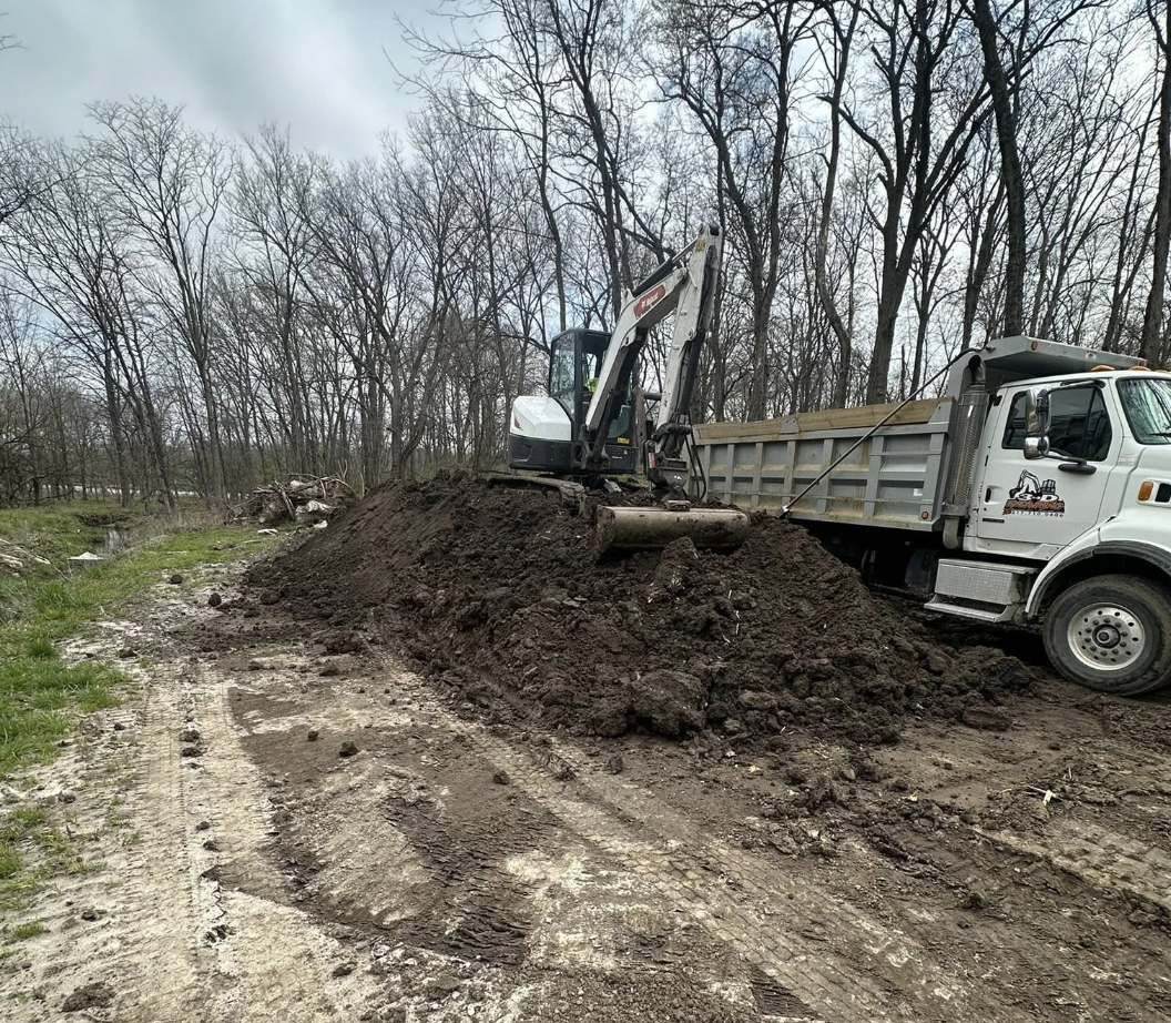 A small excavator loading dirt and soil into a dump truck at a construction site near trees, with tire tracks visible on the ground.