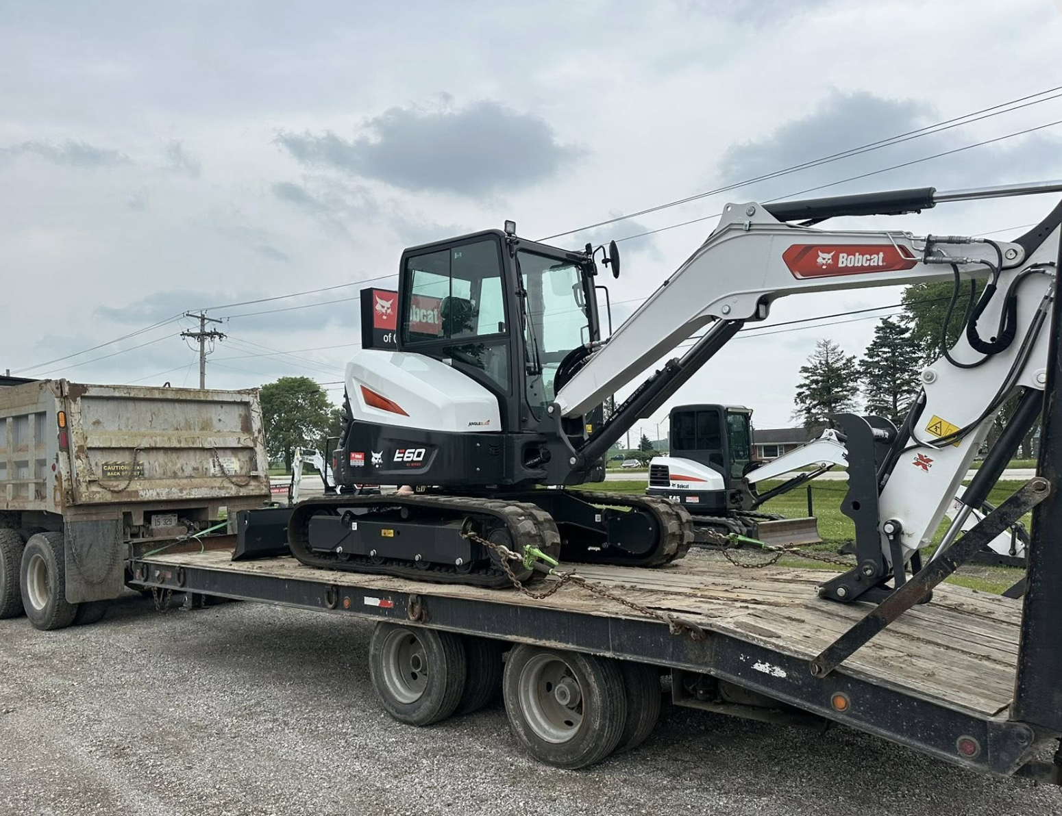 A small white and black Bobcat E60 compact excavator loaded onto a flatbed trailer, with a rusty dump truck beside it on the trailer, under cloudy skies.