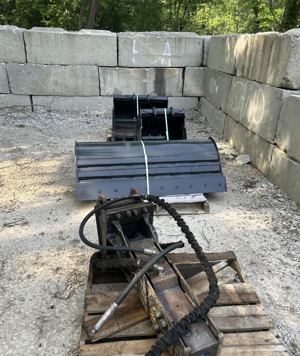 A construction site with three black excavator buckets placed on wooden pallets, situated on a gravel ground near a concrete wall with graffiti and a wooded area in the background.