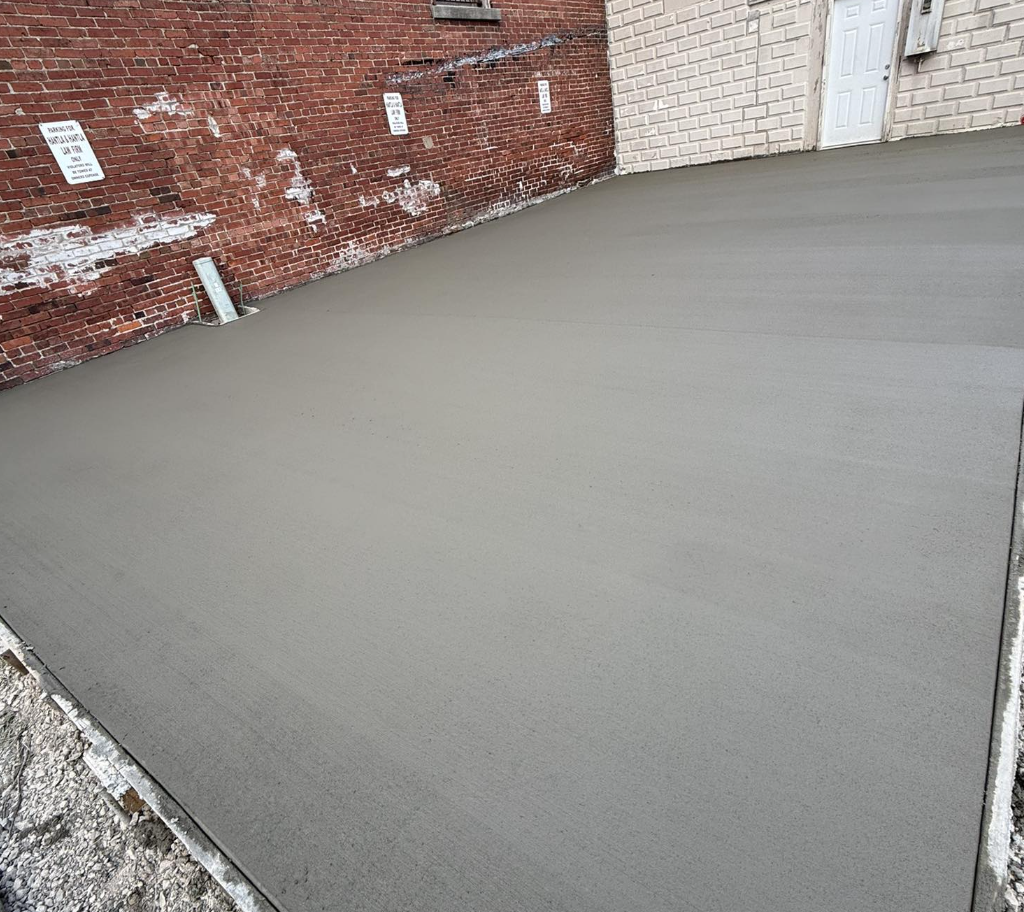 Freshly poured concrete slab in an outdoor urban area, bordered by a brick wall and a building with a white door.