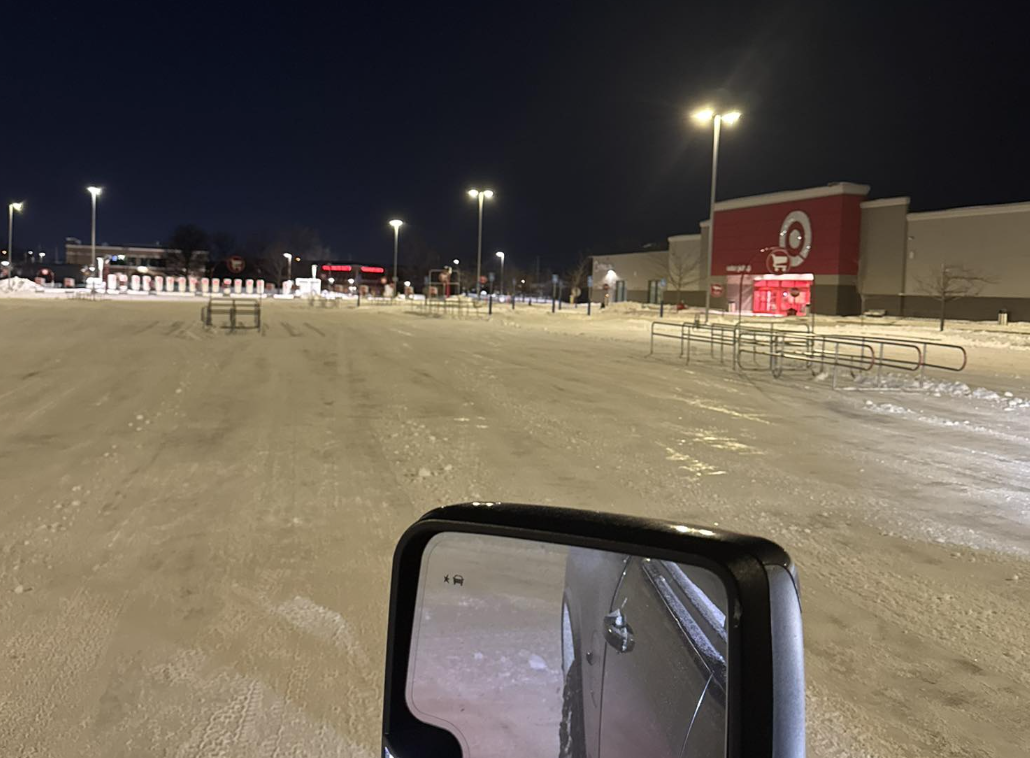 Empty parking lot at night with snow and a Target store visible in the background. A vehicle's side mirror is in the foreground.