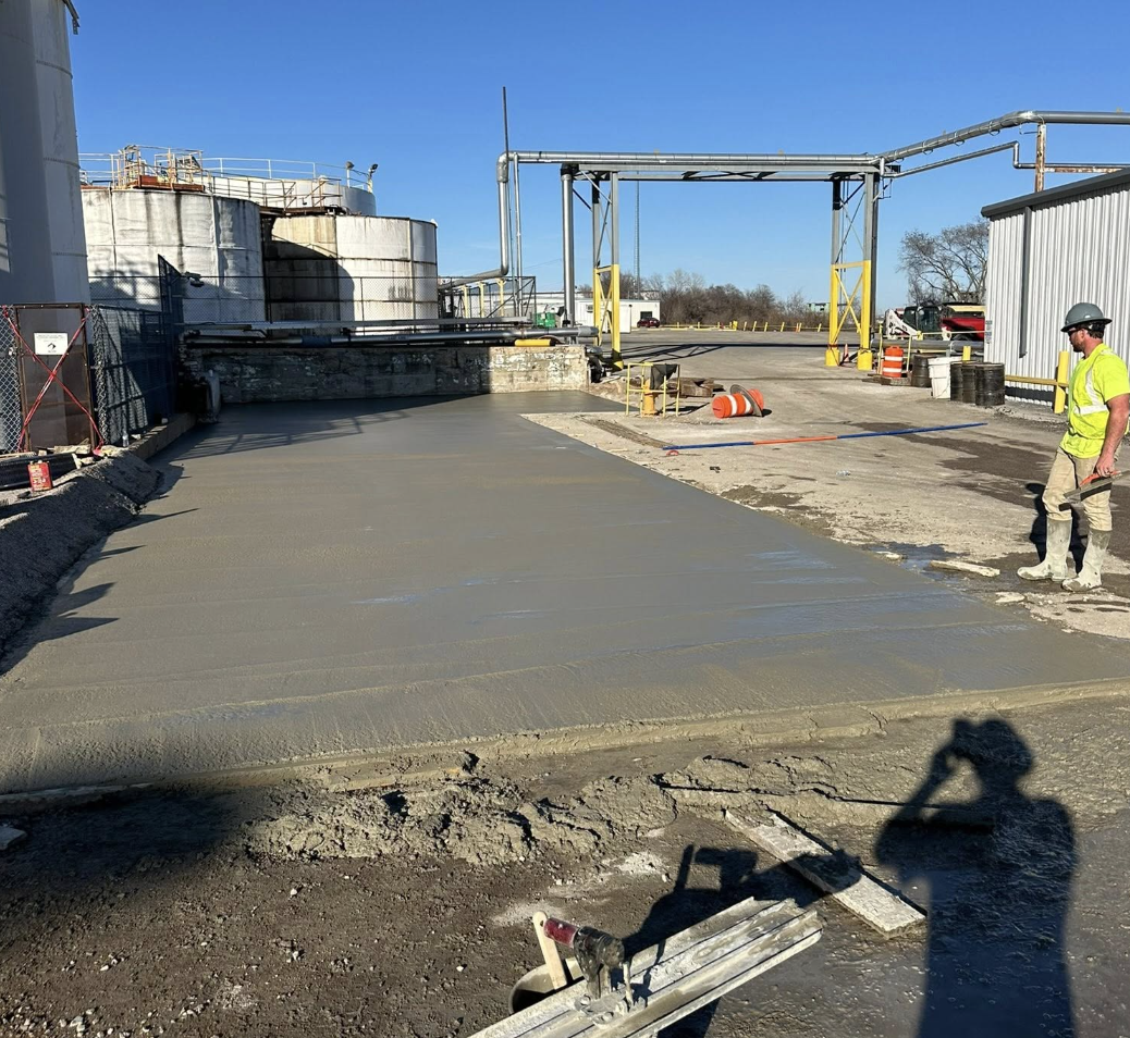 Construction worker in a yellow safety vest and helmet smoothing fresh concrete on a newly poured driveway at an industrial site under a clear blue sky.