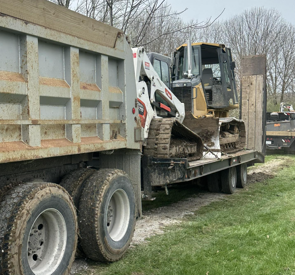 A small bulldozer loaded onto a flatbed truck with muddy tires, parked on a grassy area near some trees, with another pickup truck visible in the background.