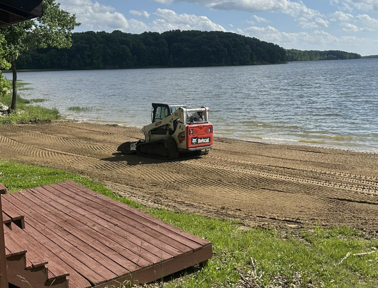A small construction vehicle, a mini skid-steer, parked on a sandy beach near a lake, with trees and hills in the background.