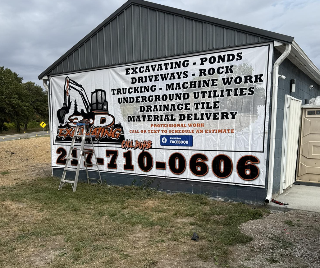 Large banner on side of a gray building advertising excavation services, including excavating, ponds, driveways, rock, trucking, machine work, underground utilities, drainage, material delivery, and professional work with a phone number 217-710-0606 