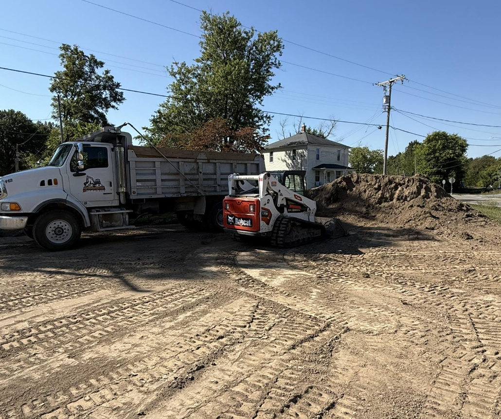 Construction site with a dump truck and a compact excavator on dirt ground, near a residential house and trees, with power lines overhead.
