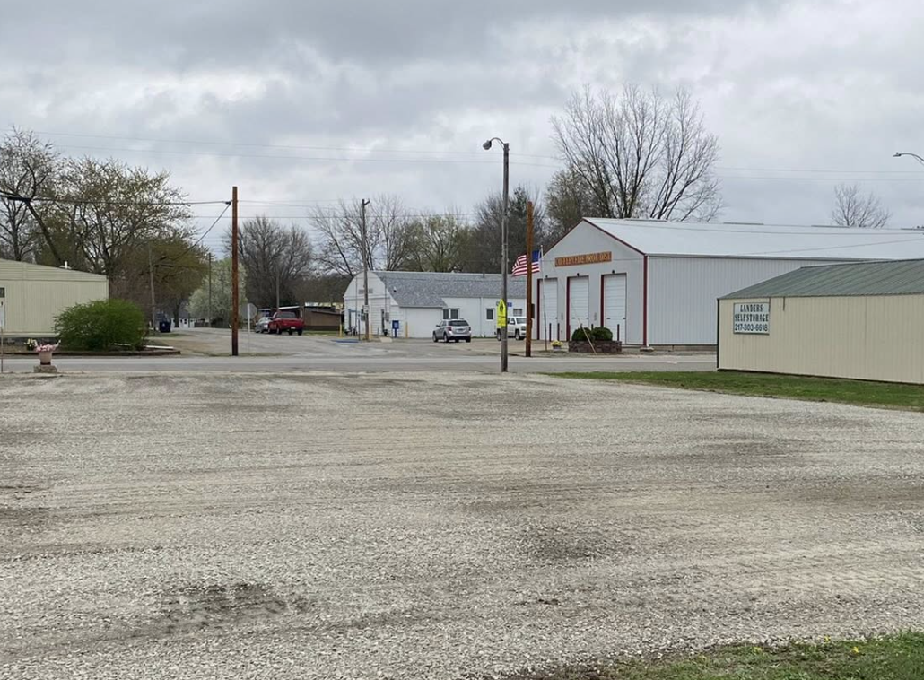 A small town street scene with gravel in the foreground, a few buildings including a fire station with an American flag, parked cars, trees, and cloudy sky in the background.