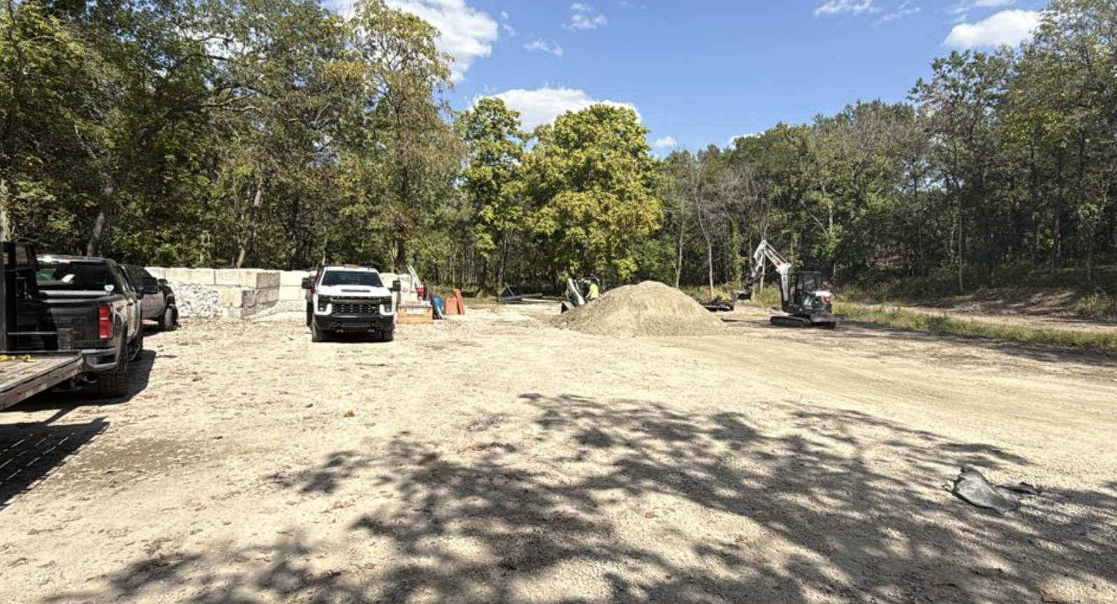 Construction site with dirt ground, parked trucks, a small excavator, and piles of dirt, surrounded by trees under a partly cloudy sky.