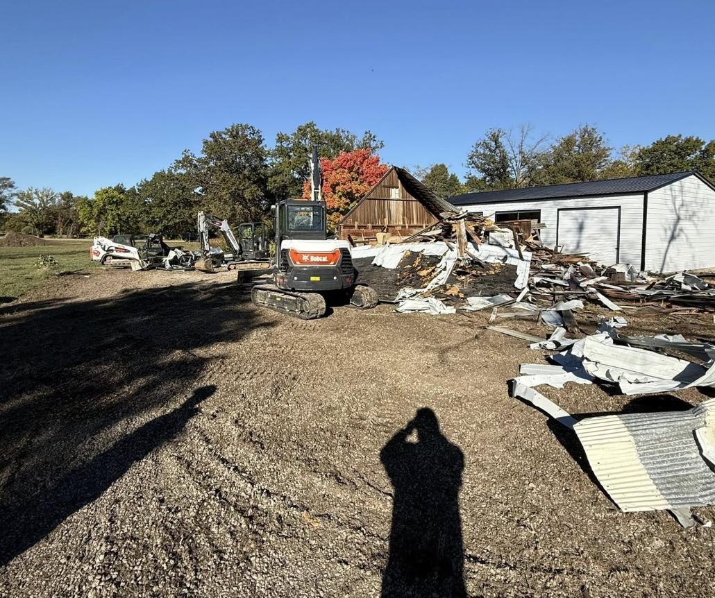 A lot of construction and demolition equipment, including a small excavator and a skid-steer loader, on a farm or rural property. There is debris and wreckage from a partially collapsed wooden structure or building. In the background, there are trees