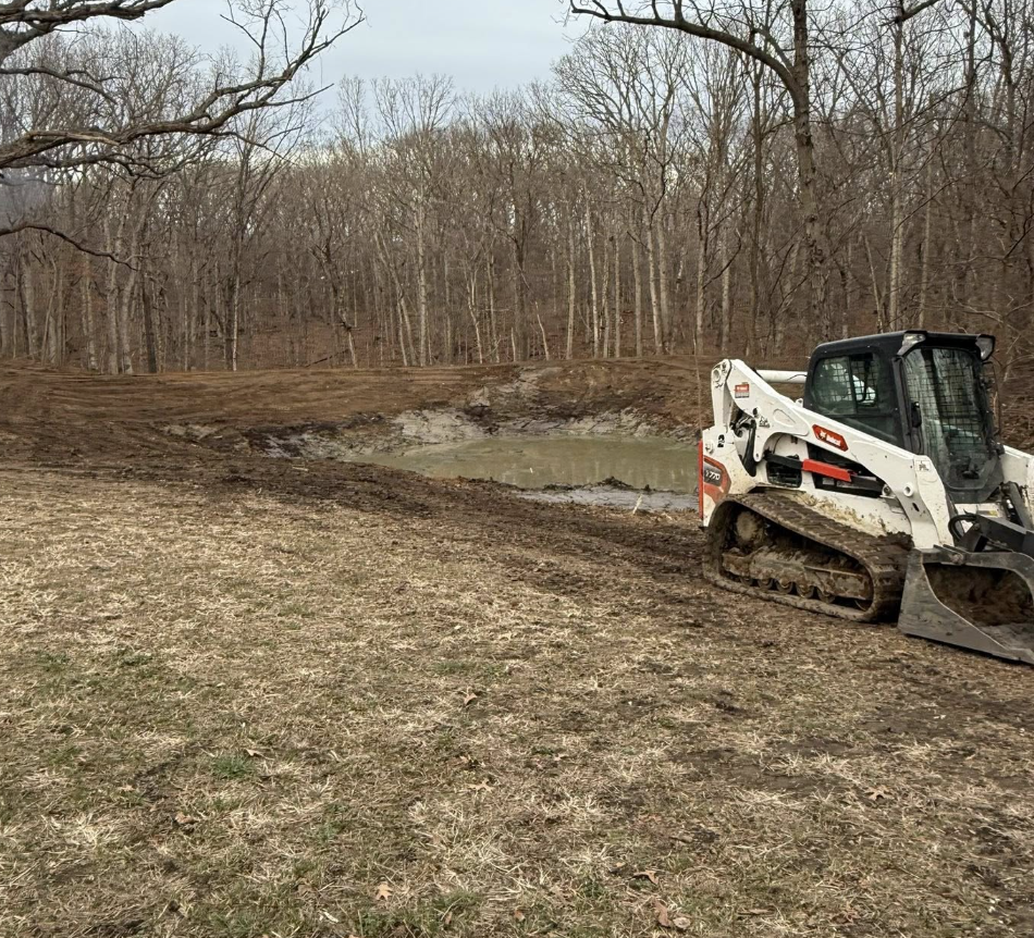 A small excavator parked on dirt ground near a muddy pond in a wooded area with leafless trees.
