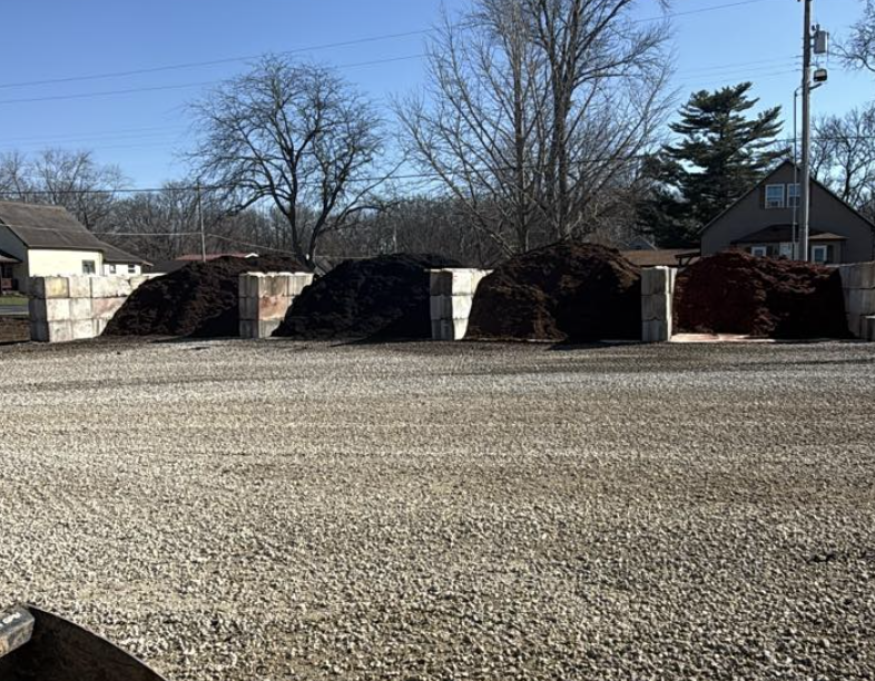 Multiple piles of dark colored gravel or soil stacked on the ground with concrete blocks behind them, set outdoors with trees and houses in the background.