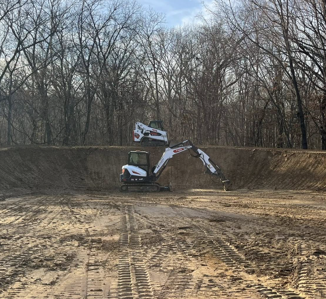 Two small excavators working on a construction site with a dirt floor, surrounded by leafless trees under a cloudy sky.