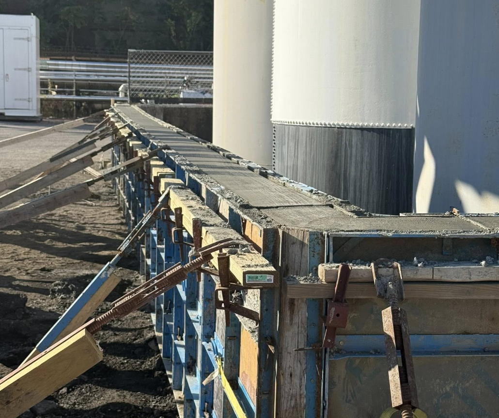 Construction site with steel formwork and clamps for pouring concrete, with large storage tanks in the background.