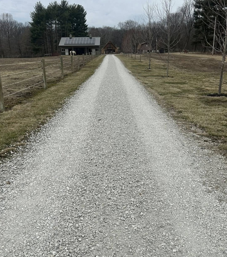 Gravel driveway leading to a house with solar panels, flanked by fenced grassy areas, with a horse in the distance and trees on either side.
